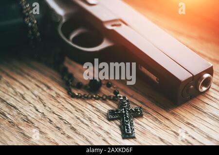 Auf einem Holztisch liegt eine Airsoft schwarze Pistole und ein schwarzes gotisches Kreuz auf einer Kette, das von einem leuchtend orangefarbenen Licht beleuchtet wird. Stockfoto