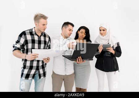 Vier junge, multiethnische Menschen, afrikanische und muslimische Mädchen, zwei kaukasische Männer, halten Papiere und verschiedene Geräte in der Nähe einer weißen Wand Stockfoto