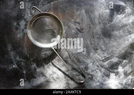 Feinmasch-Sieb aus Edelstahl, Kälte Mehlsieb mit Griff auf dunklem Hintergrund, mit Mehl bestäuben. Stockfoto