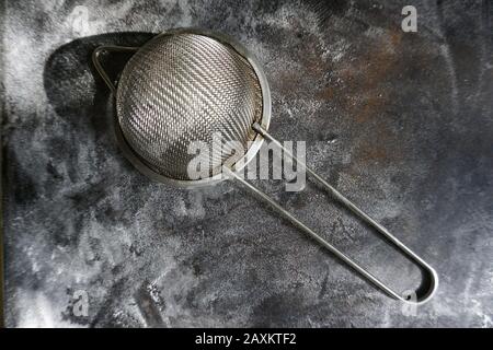 Feinmasch-Sieb aus Edelstahl, Kälte Mehlsieb mit Griff auf dunklem Hintergrund, mit Mehl bestäuben. Stockfoto
