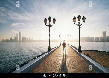 Junger Mann, der bei Sonnenaufgang auf dem Gehweg gegen das Meer und die Skyline der Stadt spazieren geht. Abu Dhabi, Vereinigte Arabische Emirate Stockfoto