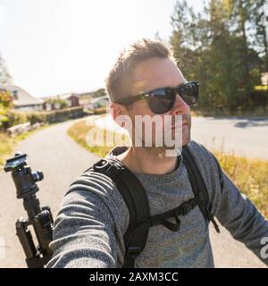 Gut aussehender, kühler kaukasischer Erwachsener mit Sonnenbrille im Freien beim gehen oder beim urbanen Wandern bei warmem, sonnigem Wetter, der einen Rucksack trägt Stockfoto
