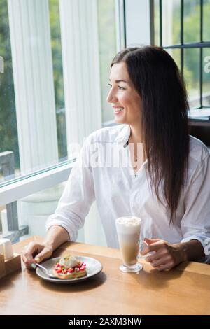 Schönen lächelnden jungen Frau im Cafe in der Nähe der Fenster mit Kaffee und leckeren Kuchen. Stockfoto