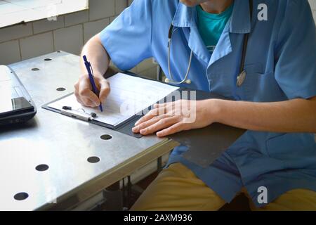 Doktor in blauer Uniform an einem Metalltisch sitzend und schreibt in einem Dokument Stockfoto