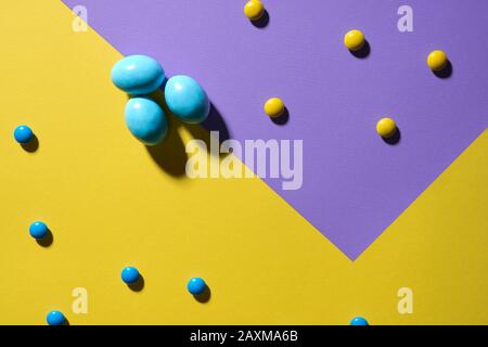 Von oben betrachtet schokolade Erdnusskugeln in bunter knuspriger Zuckerschale und drei blaue Eier isoliert auf gelbem und violett-geometrischem Hintergrund. Nahaufnahme von hübschen Süßigkeiten, die auf dem Tisch liegen, Schatten. Stockfoto