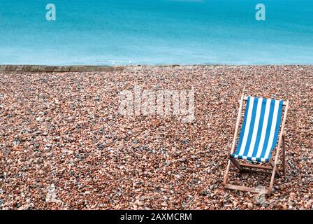 Ein verlasser Kieselstrand mit einem leeren gestreiften Liegestuhl unten rechts im Vordergrund. Zwei Möwen sitzen auf einer Steingroyne im Hintergrund mit dem Meer Stockfoto