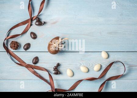Dunkle und weiße Schokolade in Form von Eiern und Kaninchen mit braunem Band auf blauem Holzhintergrund. Festliche Grußkarte für den Osterfrühling. Draufsicht Stockfoto