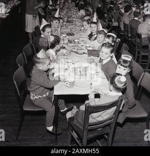 1960er Jahre, historisch, in einem Speisesaal, kleine Schulkinder in Partyhüten, die gemeinsam bei einem weihnachtsessen Spaß haben, England, Großbritannien Stockfoto