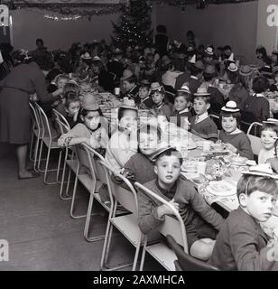 1960er Jahre, historisch, in einem Speisesaal, kleine Schulkinder in Partyhüten, die gemeinsam bei einem weihnachtsessen Spaß haben, England, Großbritannien. Stockfoto