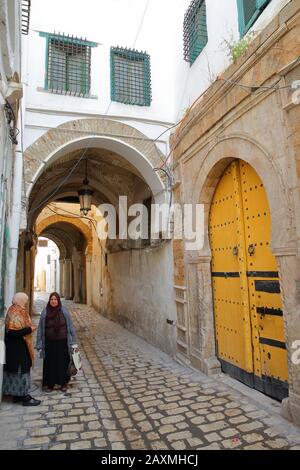 Tunis, TUNISIA - 30. DEZEMBER 2019: Typische gepflasterte und enge Straße mit Arkaden in der historischen Medina von Tunis und zwei einheimischen Frauen Stockfoto