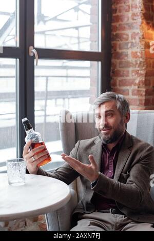 Aufgeregter gutaussehender grauhaariger Mann, der in einem luxuriösen Sessel sitzt und Whisky einem Freund empfiehlt Stockfoto