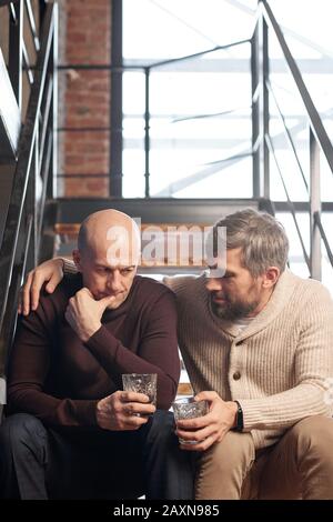 Bärtiger Mann im mittleren Alter in Strickjacke auf der Treppe sitzend und dem engen Freund beim Alkoholtrinken Ratschläge geben Stockfoto