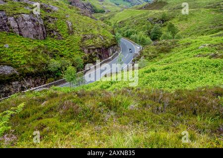 Ein einzelnes Motorrad, das im Sommer durch ein üppiges Tal in den schottischen Highlands Scotland durch eine Kurve auf der Landstraße fährt Stockfoto