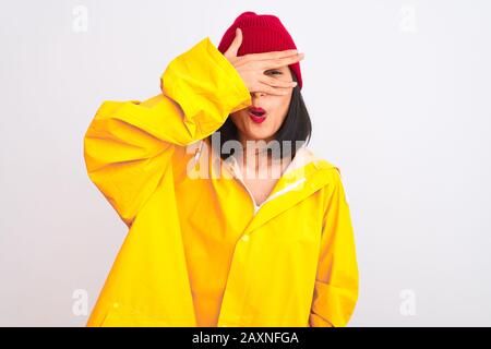 Junge, schöne chinesin, die Regenmantel und Wollkappe über isoliertem weißem Hintergrund trägt und dabei mit der Hand Gesicht und Augen bedeckt, sieht es aus Stockfoto