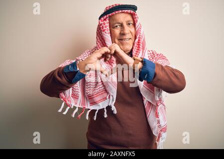 Ein hochrangiger, gutaussehender arabischer Mann, der Keffiyeh trägt, der über einem isolierten weißen Hintergrund steht und in Liebe lächelt, um Herzsymbol und Form mit den Händen zu zeigen. Romantisch Stockfoto