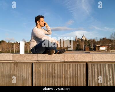 Junger gutaussehender kaukasischer Mann, der am Telefon spricht und in Madrid Rio mit der Kathedrale von Almudena hinter sich sitzt. Madrid, Spanien. Stockfoto