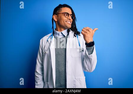 Junger afroamerikanischer Arzt mit Dreadlocks, die Stethoskop und Brille tragen und mit einem fröhlichen Gesicht lächeln, das mit dem Daumen auf die Seite zeigt Stockfoto