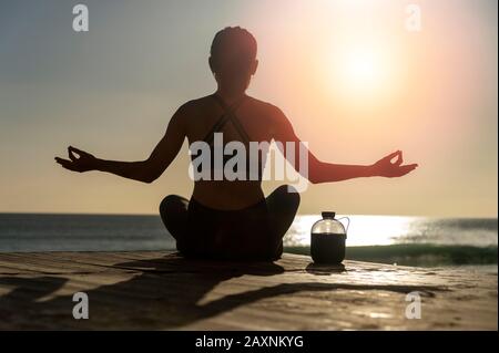 Silhouette einer Frau, die bei Sonnenaufgang oder Sonnenuntergang am Meer meditiert, Wasserflasche. Stockfoto