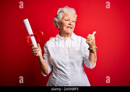 Ältere, schöne Studentin mit Diplom, die über einem isolierten roten Hintergrund steht und mit Daumen nach oben zur Seite zeigt, mit glücklichem Gesicht sm Stockfoto