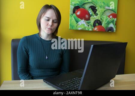 Mädchen mit einem skeptisch-nachdenklichen Gesichtsausdruck sitzt an einem Laptop an einem Tisch in einem Café auf einem braunen Sofa gegen eine gelbe Wand. Stockfoto