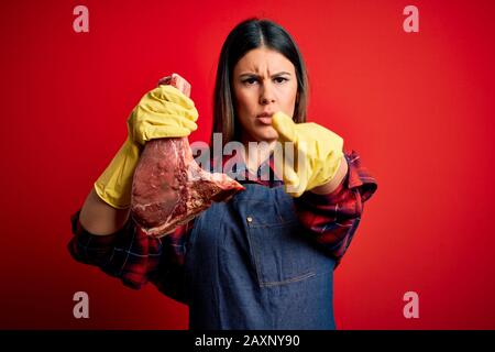 Junge Metzgerin hält frischen rohen Fleischpfahl über rotem Hintergrund und zeigt mit dem Finger auf die Kamera und auf Sie, Handzeichen, positiv und konfi Stockfoto