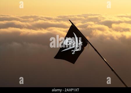 Jolly Roger (Piratenfahne) auf Wind und Meer bei Sonnenuntergang bewölkt Himmel im Hintergrund Stockfoto