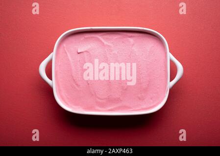 Hausgemachtes Himbeereis in einem weißen Keramikbehälter auf einem üppigen, lavafarbenen Tisch. Fruchtiges Eis. Köstliche rote Beeren Gelato. Sommerdessert. Stockfoto