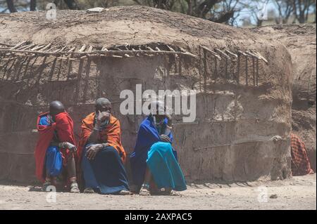 Drei Masai-Frauen sitzen vor einem traditionellen Dunghaus und schützen ihre Augen vor dem Staub. Stockfoto