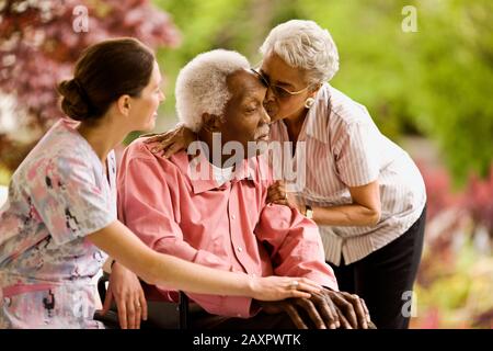 Die Krankenschwester legt die unterstützende Hand auf das Knie eines älteren Mannes, während sich eine ältere Frau auflehnt, um ihn zu umarmen und seine Stirn zu küssen. Stockfoto