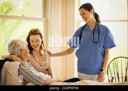 Die Krankenschwester legt eine beruhigende Hand auf die Schulter einer jungen Frau, als sie ihren Arm um eine ältere Frau legt, die in einem Bett sitzt. Stockfoto