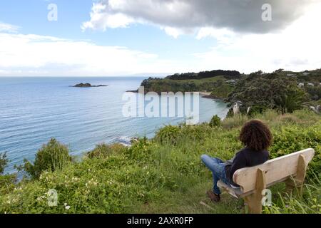 Mann mit lockigen Haaren, der auf das Meer blickt, sitzt auf einer Bank, ist Waiheke. Stockfoto