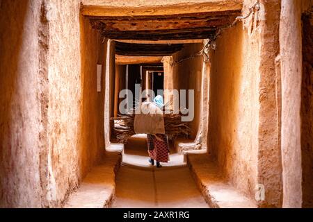 El Khorbat, Marokko - Frau, die eine Last trockener Palmblätter durch die schmale Gasse trägt Stockfoto