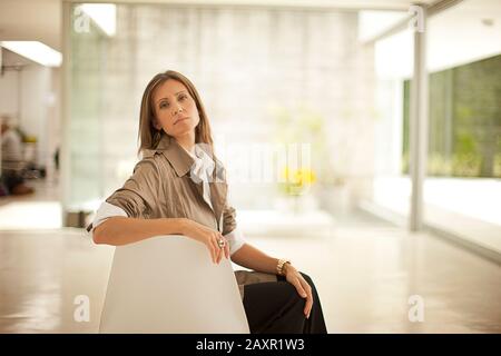 Geschäftsfrau posiert selbstbewusst für ein Porträt, als sie vor großen Fenstern im Foyer eines Büros sitzt. Stockfoto