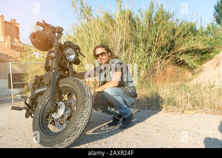 Langhaariger Brunette-Typ in Sonnenbrille Jeans und ein Jeanshemd, das auf einem schwarzen, individuellen Motorrad posiert Stockfoto