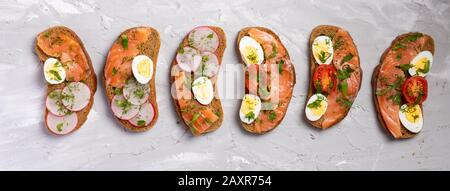 Geröstetes Roggenbrot mit Räucherlachs, Rettich und Quial-Eiern. Köstlicher gesunder Snack mit frischem grünen Gemüse auf grauem Tisch, Fußtritten von oben Stockfoto