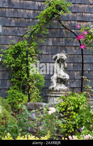 Skulptur des Jungen mit Delphin und schwarzem Schmiedeeisen Arbour bedeckt mit Hedera - Klettern Ivy neben alt Verwittertes Zedernschindelgebäude Stockfoto