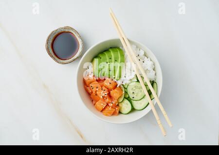 Lachs-Poke-Schüssel mit frischem Fisch, Reis, Gurke, Avocado mit schwarzem und weißem Sesam. Weißer Marmortisch. Lebensmittelkonzept. Stockfoto