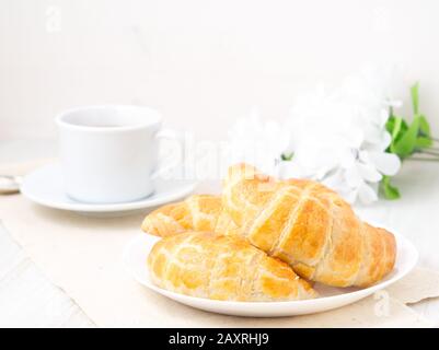 Tasse Tee, Blumen und frisch gebackenes Blätterteig auf einem weißen Tisch. Köstliche Croissants mit seitlichem Blick Stockfoto