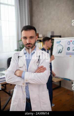 Bärtiger junger Arzt, der mit gekreuzten Armen steht Stockfoto