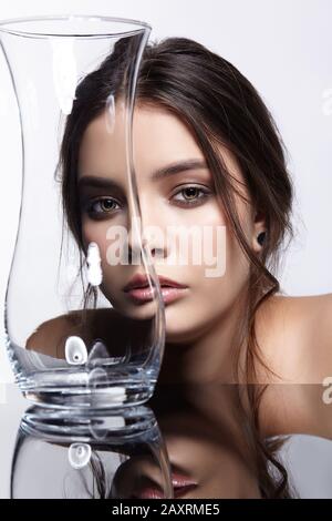 Mädchen verbirgt ihr Gesicht hinter einem Glas Vase. Schönheit Porträt der jungen Frau im Spiegel Tisch. Weibliche auf grauem Hintergrund. Stockfoto