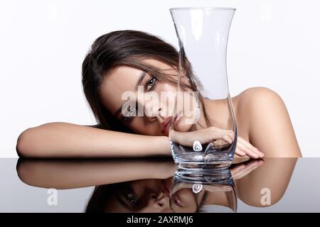 Mädchen verbirgt ihr Gesicht hinter einem Glas Vase. Schönheit Porträt der jungen Frau im Spiegel Tisch. Weibliche auf grauem Hintergrund. Stockfoto