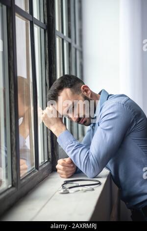 Junger bärtiger Mann, der in Gedanken in der Nähe des Fensters steht Stockfoto