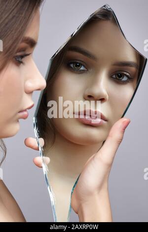 Mädchen mit einem Schard des Spiegels. Weibchen mit Spiegelschard in der Hand, die auf grauem Hintergrund posiert. Gesichtsreflexion im Spiegelsplitter. Stockfoto