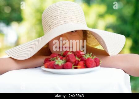 Portrait des Fröhlichen netten Mädchen in großer Mütze isst im Sommer Erdbeeren Stockfoto