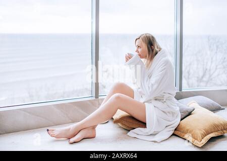 Frau in einem weißen bademantel mit Meerblick neben einem großen Fenster mit einer Tasse Kaffee. Panoramafenster. Bodenbelag, Urlaub in einem Hotel Stockfoto