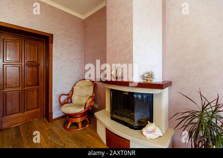 Elektrischer Kamin im Zimmer, in der Ecke befindet sich ein Korbstuhl, die richtige Anlage. Modelle von Segelbooten an den Mantelstücken. Beige Wände Stockfoto