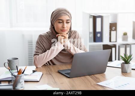 Eine nachdenkliche muslimische Geschäftsfrau, die am Arbeitsplatz sitzt und einen Laptop-Bildschirm betrachtet Stockfoto