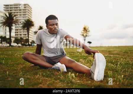 Ein junger afroamerikanischer Fitness-Mann, der vor dem Workout auf grünem Gras sitzt und seine Beine streckt Stockfoto