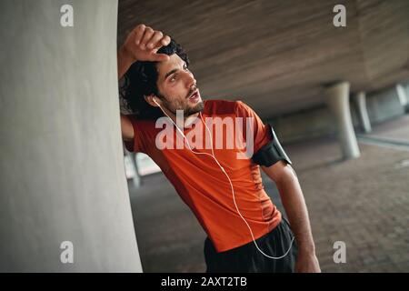 Erschöpfter sportlicher junger Mann mit Ohrhörern und Armband, der sich an die Säule unter der Brücke lehnt und sich nach dem laufen ausruhen kann - erschöpfter Athlet Stockfoto