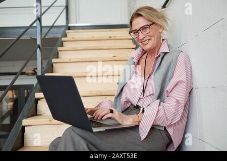 Porträt einer fröhlichen, reifen Geschäftsfrau, die auf einer Treppe sitzt und einen Laptop verwendet, während sie den Verkaufsbericht vorbereitet Stockfoto